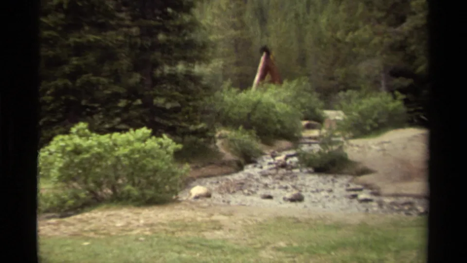 Canadian 1979 8mm frame showing a distant walker in red moving behind shrubs near rocks, watercourse, grass, and evergreens.
