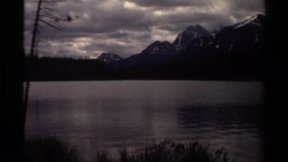 1979 Canadian 8mm frame showing calm lake water, distant snow-capped mountains, and a cloudy sky.