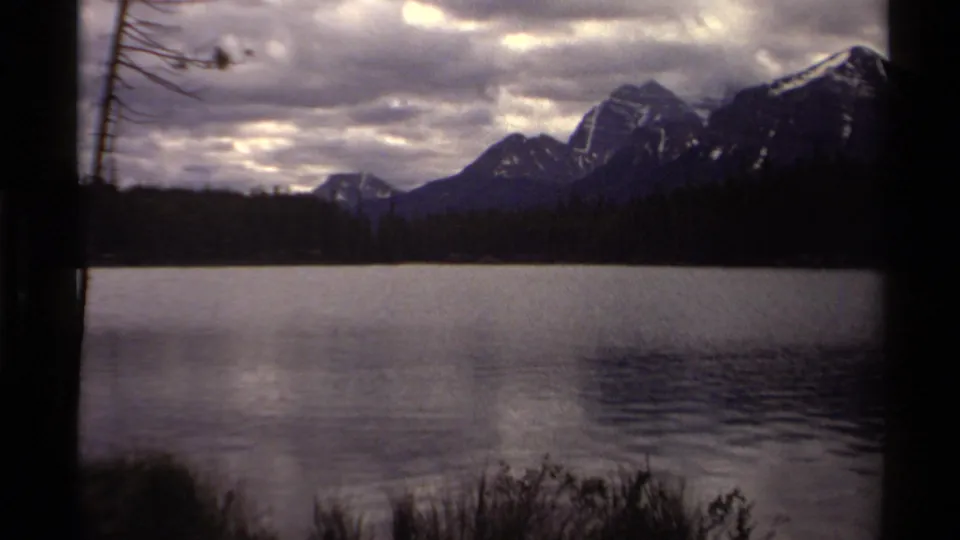 1979 Canadian 8mm frame of still lake water with distant snow-capped mountains and muted cloud reflections.