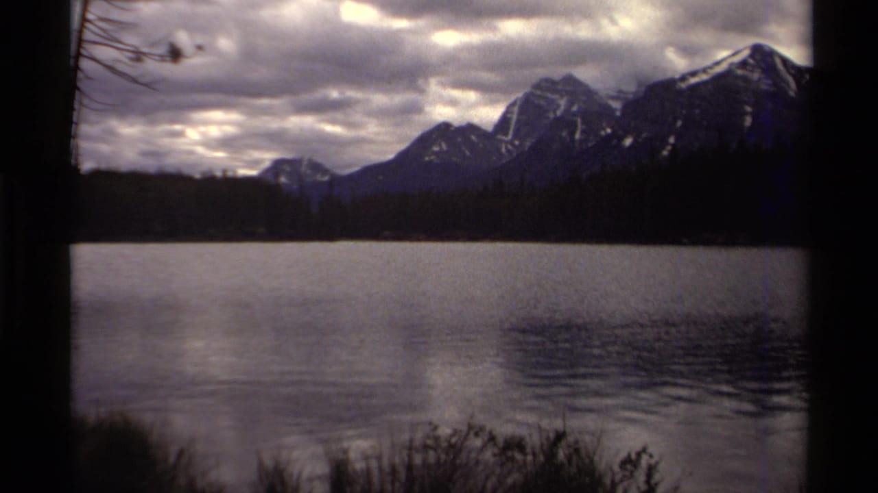 1979 8mm home-movie view in Canada of a lake, dark foreground trunk, pine shoreline, and mountains beneath heavy clouds.