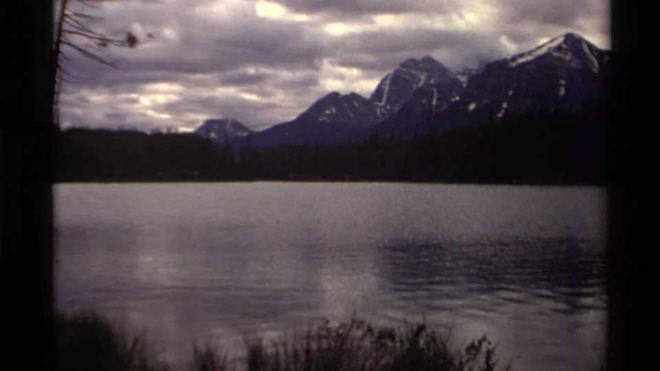 1979 8mm home-movie view in Canada of a lake, dark foreground trunk, pine shoreline, and mountains beneath heavy clouds.