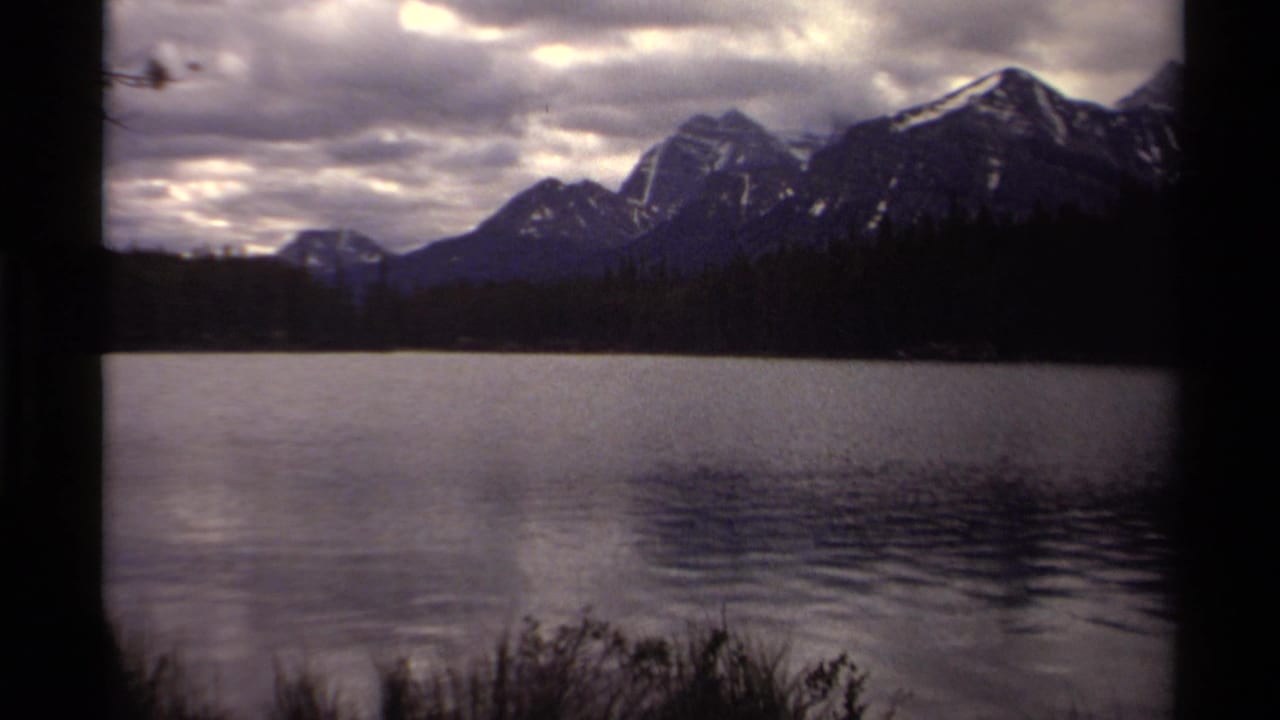 Canadian mountain lake scene on 8mm film, with pine forest, dark vertical foreground framing, and snow-streaked peaks.