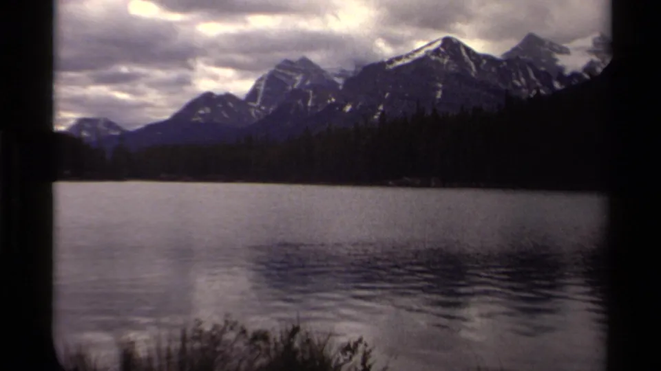 1979 Canadian 8mm frame showing a lake, wooded shoreline, and snow-streaked mountains under a layered cloudy sky.