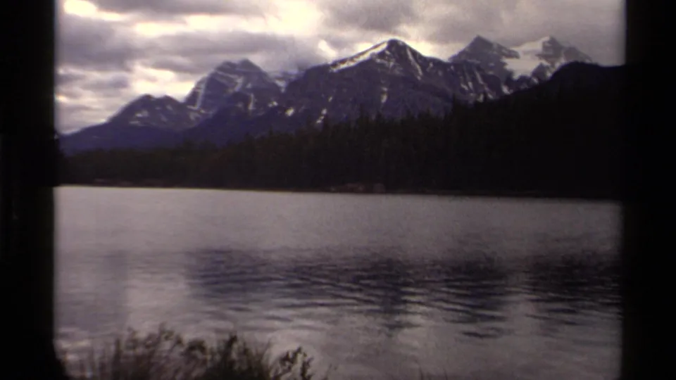 Vintage 8mm view of a Canadian lake bordered by pines, with mountains and a dark foreground trunk under overcast sky.