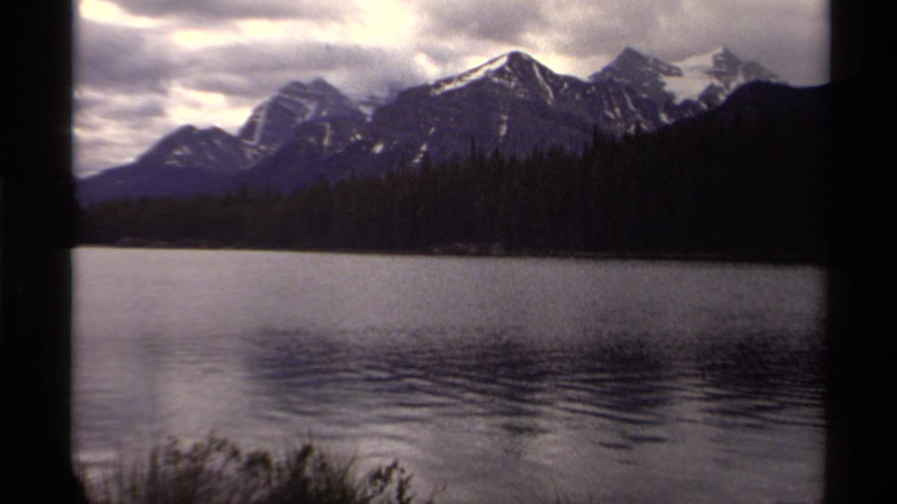Snow-capped mountains behind a dark forest and calm lake under cloudy sky.