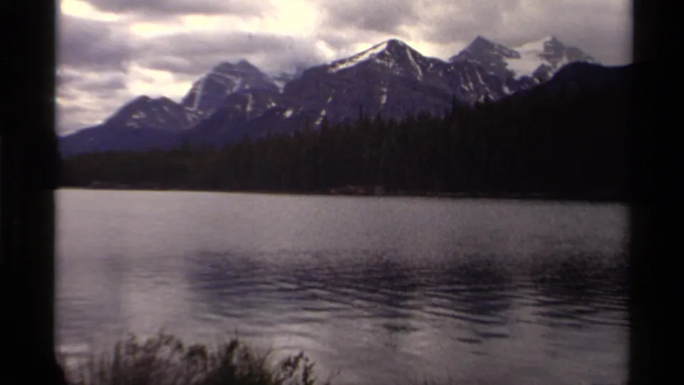 1979 Canadian 8mm frame of a mountain lake with snowy peaks, evergreen forest, rippled water, and overcast sky.