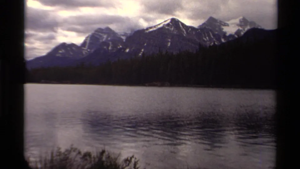Canadian 1979 home-movie view of snowy mountain peaks above a dark forest and broad lake surface.
