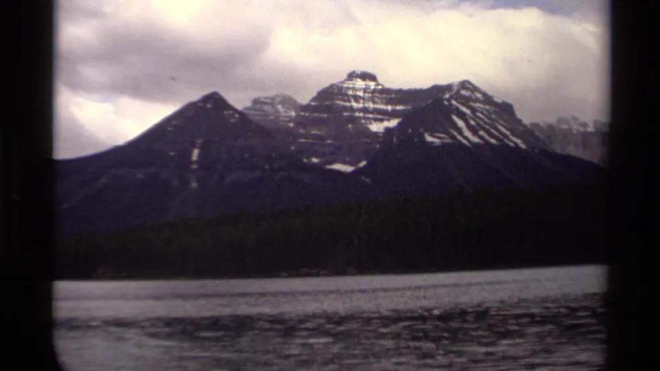 1979 8mm Canadian landscape frame showing layered mountains, snow bands, forest, lake edge, and cloudy light.