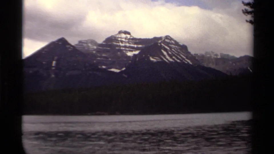 1979 Canadian 8mm frame with snow-capped peaks, shadowed forest, rippling lake water, and overcast sky.
