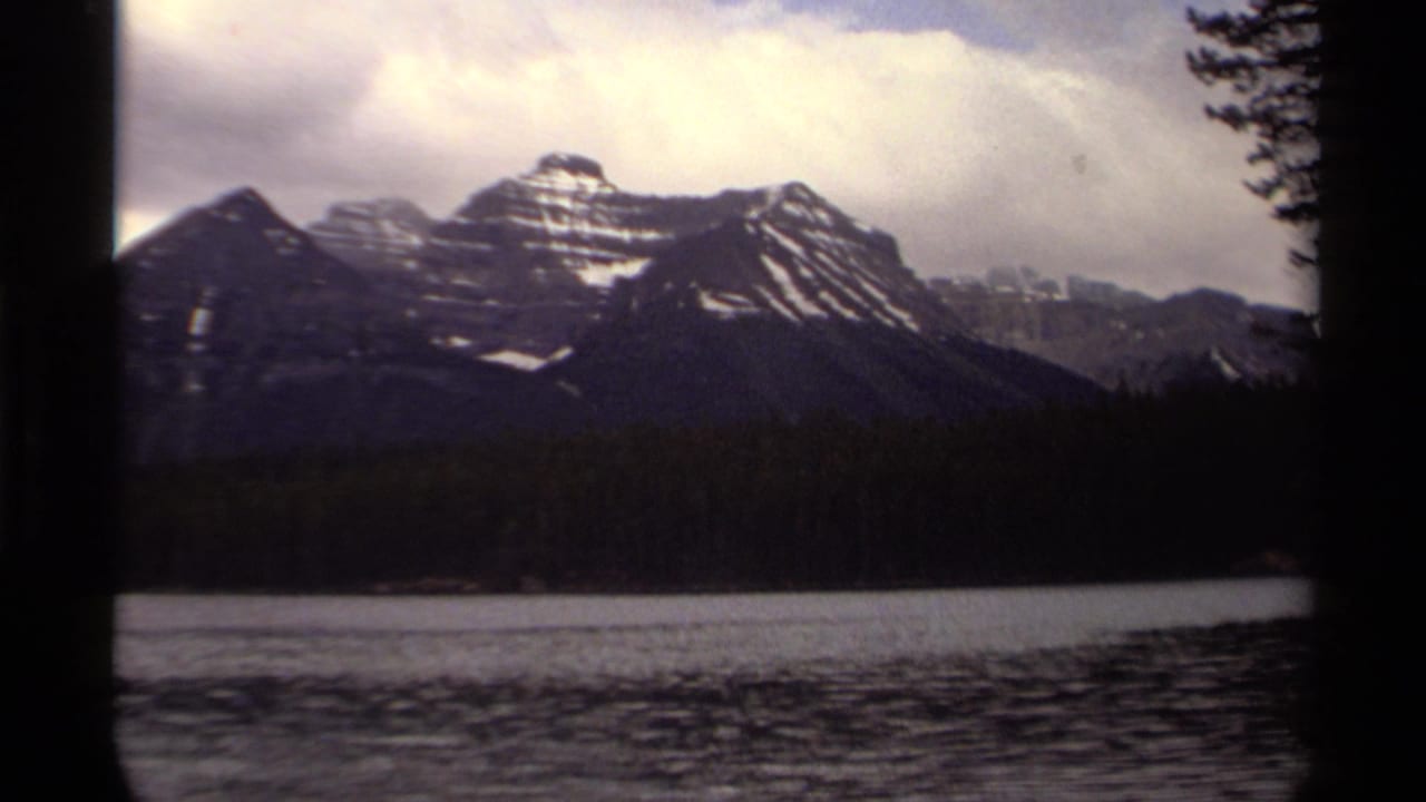 1979 8mm Canadian home-movie frame showing a calm lake, evergreen shoreline, and snow-dusted mountains beneath clouds.