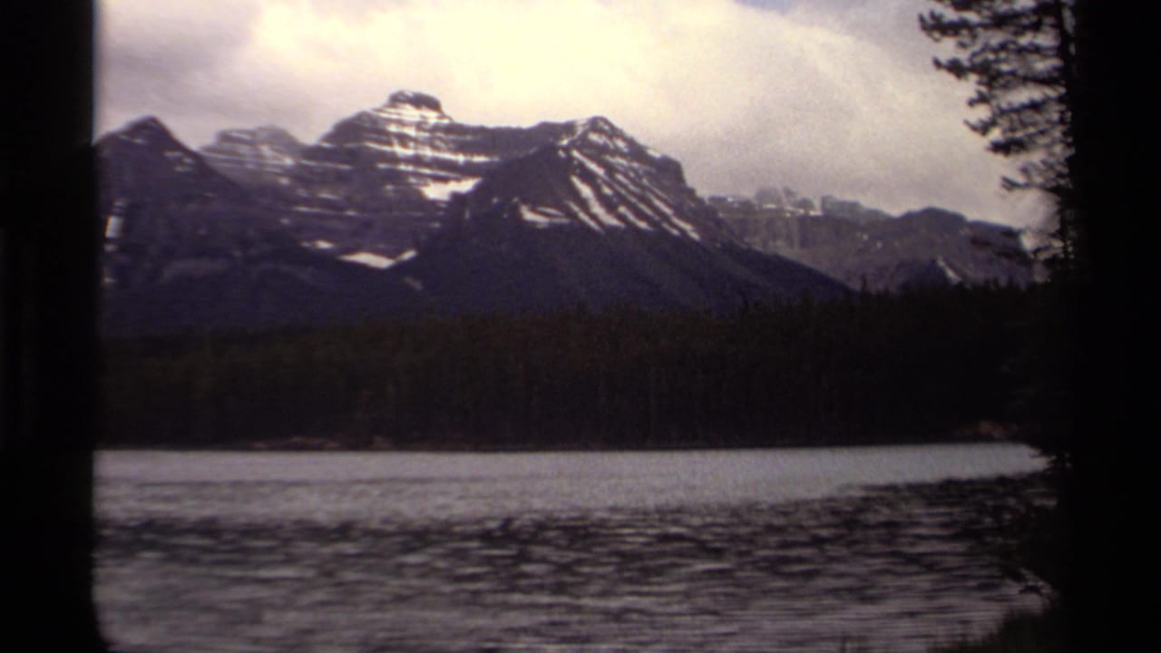 Canadian 8mm home-movie view of snow-streaked peaks, dark evergreen forest, and a calm lake surface framed by trees.