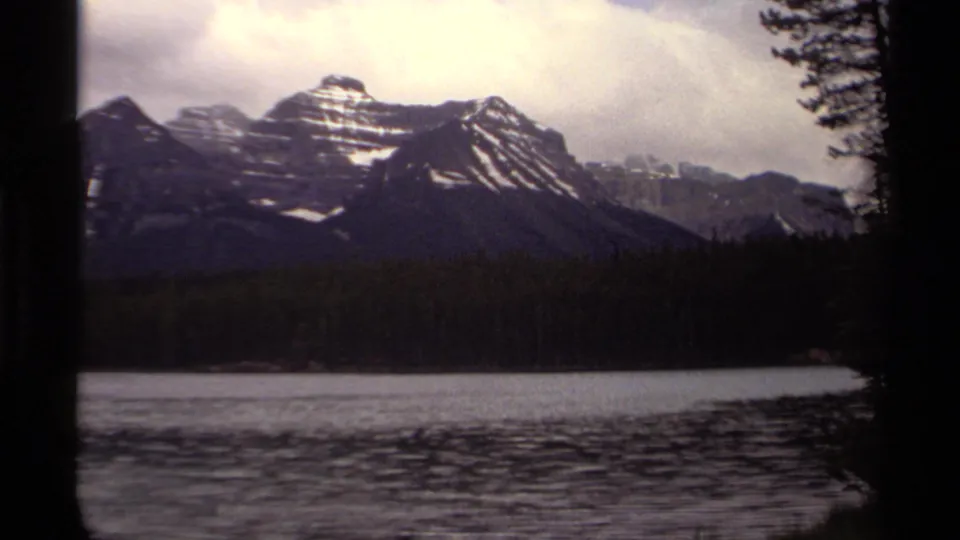 Canadian 8mm home-movie view of snow-streaked peaks, dark evergreen forest, and a calm lake surface framed by trees.