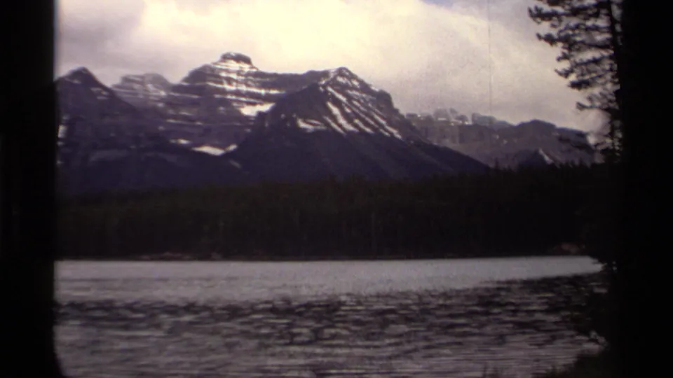 1979 8mm Canadian landscape frame with bright cloud cover over mountains, evergreen shore, and gently rippled lake water.