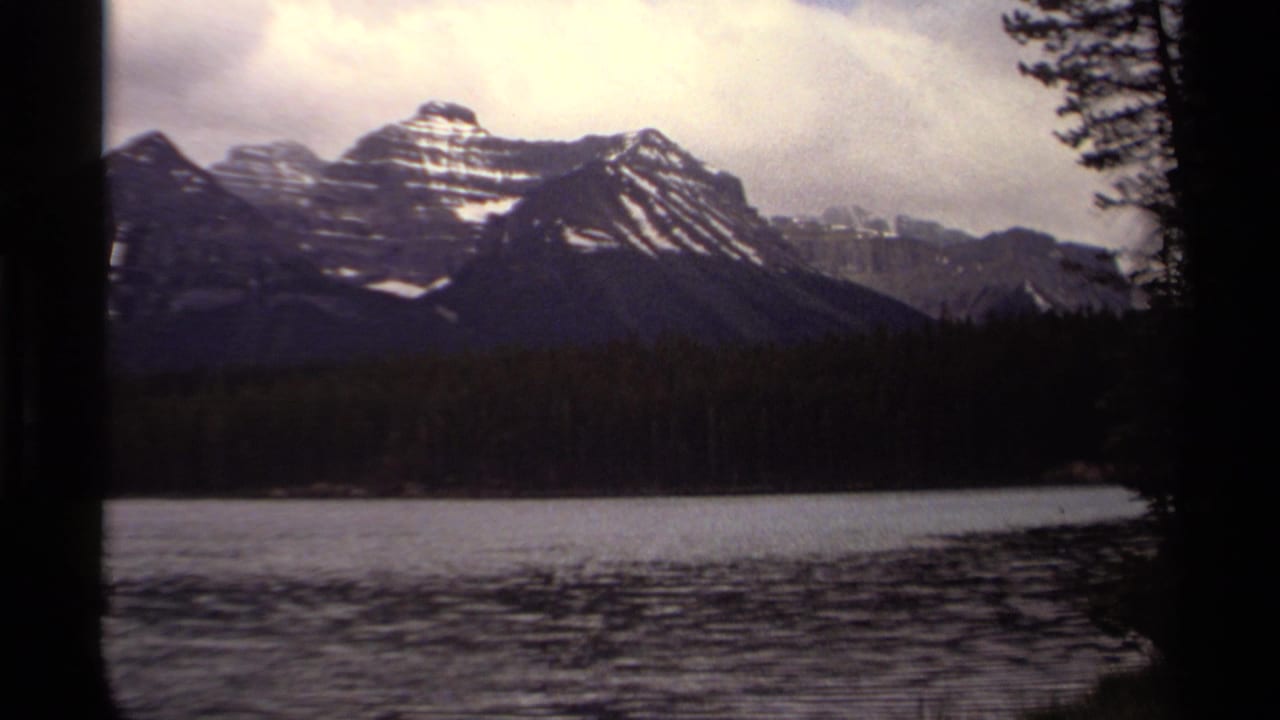 1979 Canadian 8mm frame showing distant snow-capped mountains, dark shoreline trees, calm lake water, and an overcast sky.