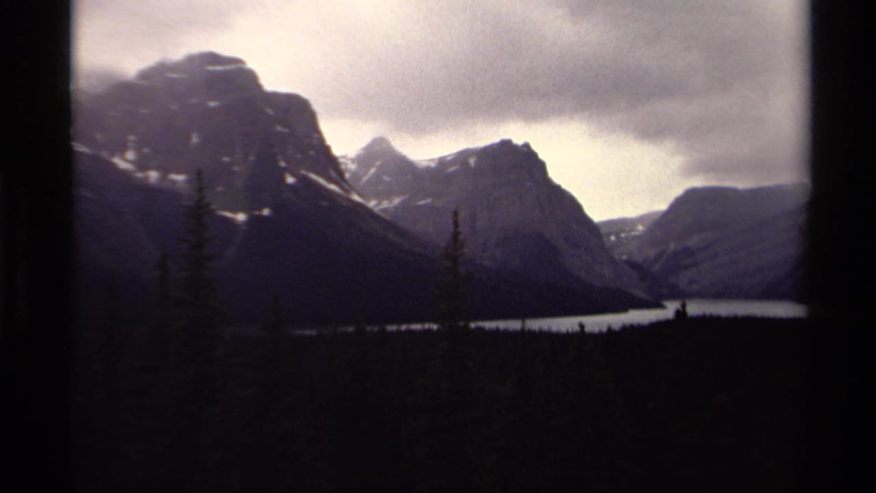 1979 Canadian 8mm view of a mountain lake bordered by evergreen forest and shadowed peaks under heavy clouds.