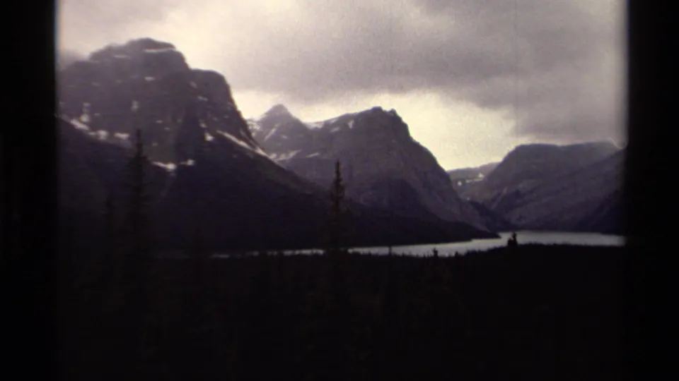 1979 Canadian 8mm frame of a broad lake, dark conifer forest, and layered mountain ridges under a dim sky.