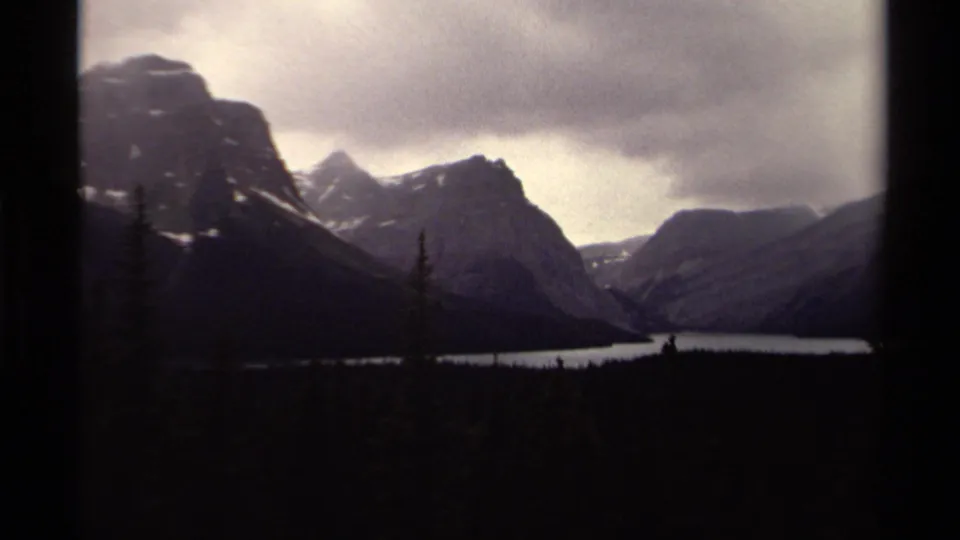 1979 Canadian 8mm frame showing a mountain lake, dark tree line, snow-marked peaks, and low overcast clouds.