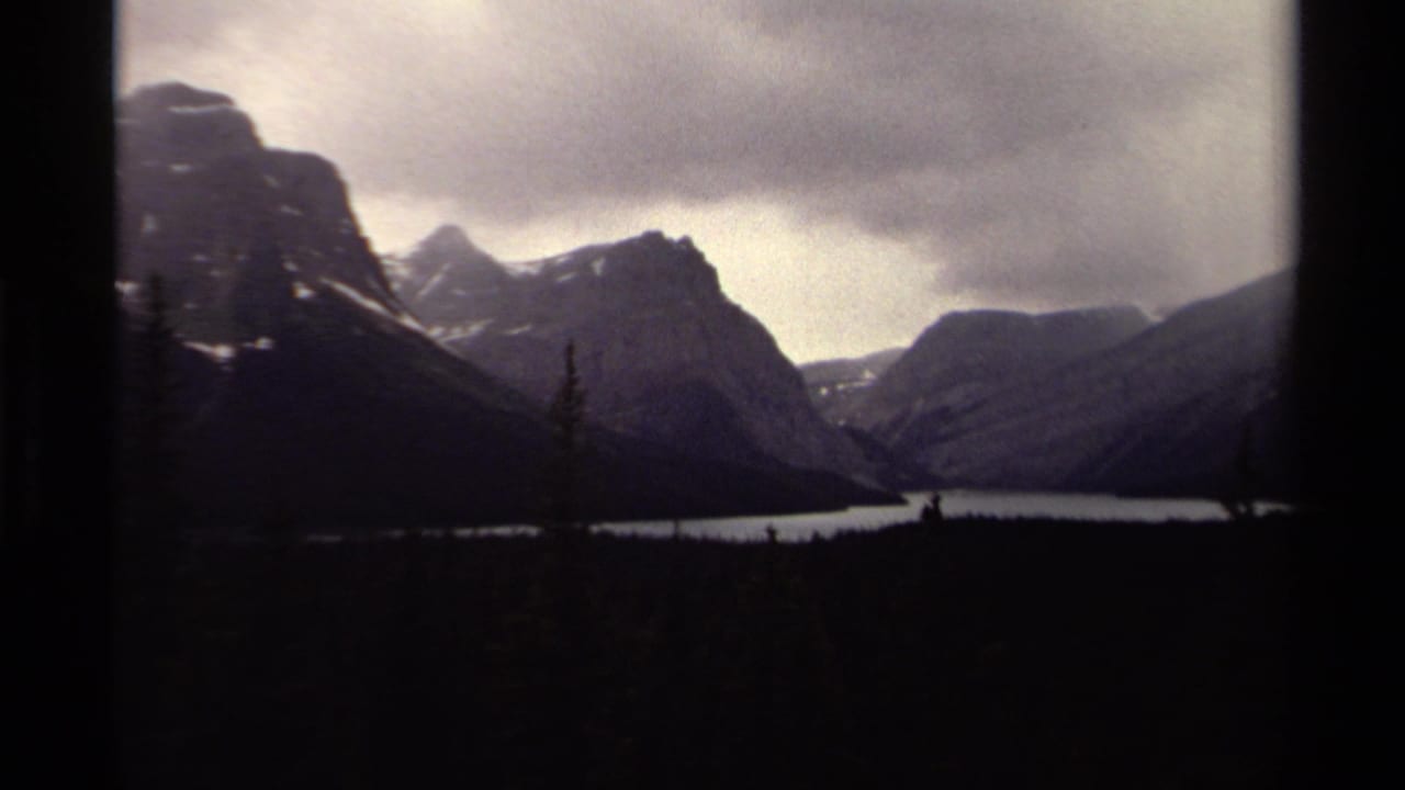1979 Canadian 8mm frame showing a dark mountain valley, pale lake, evergreen silhouettes, and low cloud cover.
