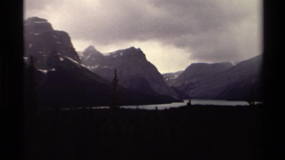1979 Canadian 8mm frame showing a dark mountain valley, pale lake, evergreen silhouettes, and low cloud cover.