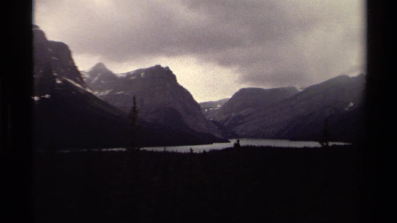 1979 Canadian 8mm view of mountain peaks above a calm lake, with dark evergreen foreground and visible frame edge.