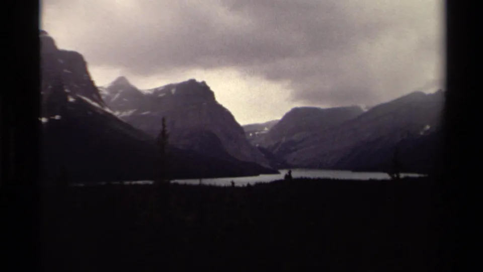 1979 Canadian 8mm view of mountain peaks above a calm lake, with dark evergreen foreground and visible frame edge.