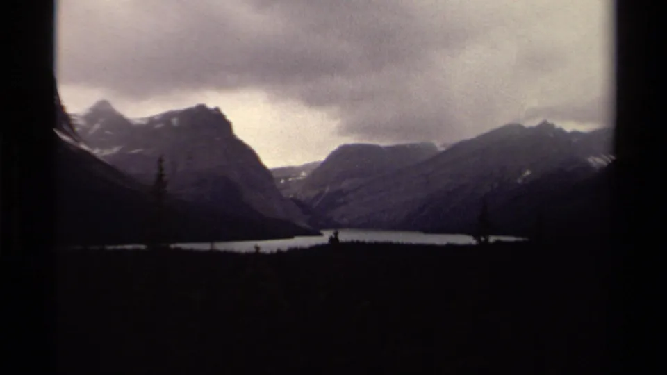 1979 Canadian 8mm frame with distant mountain ridges, a pale lake, silhouetted evergreens, and heavy clouds.