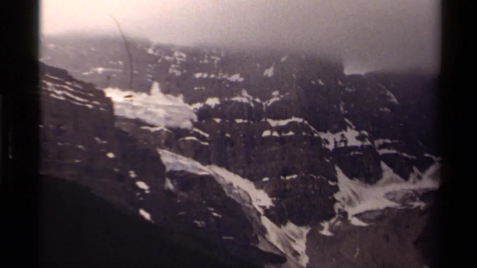 Vintage 8mm frame of a Canadian mountain landscape with layered cliffs, scattered snowfields, and low cloud cover.