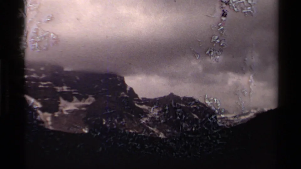 1979 8mm Canada frame of cloudy snow-patched mountains with pronounced film surface marks and gate-edge shadow.