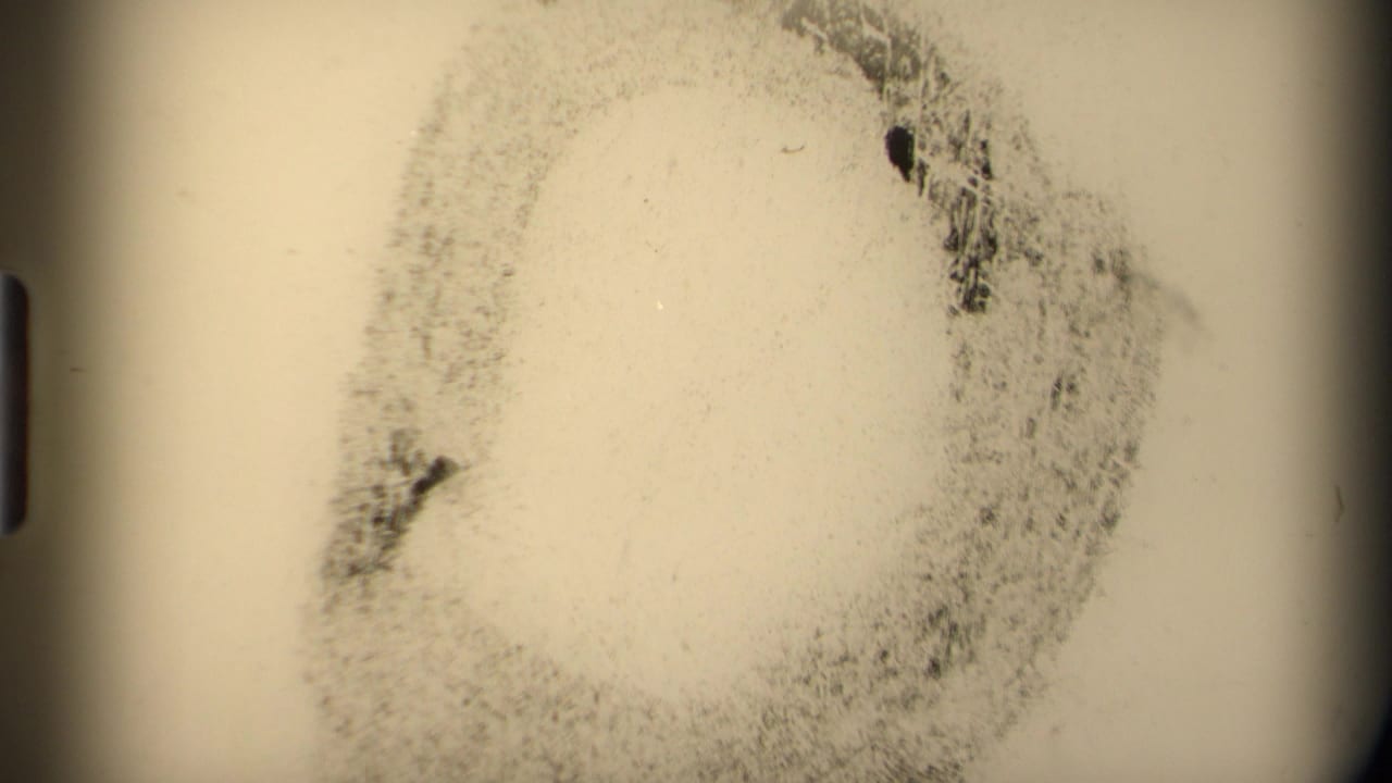 Pale 8mm frame with mottled grain, a circular dark mark, and rounded black frame edges, likely a film artifact or transitional exposure.