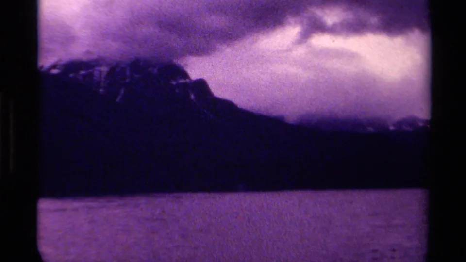 1979 8mm Canada frame showing a dark mountain ridge, cloudy sky, and water in the foreground with heavy grain and purple color shift.