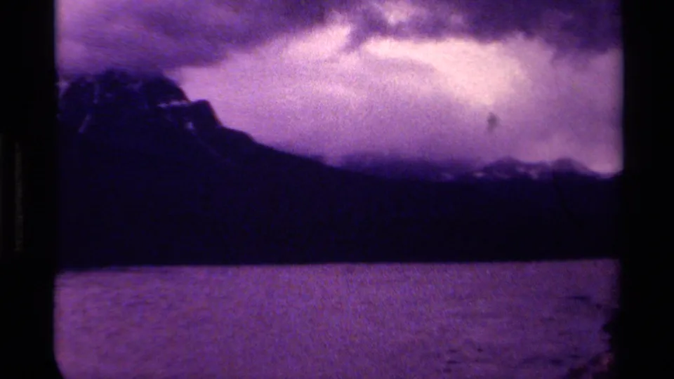 Canadian 1979 8mm frame with mountain slope, water foreground, low clouds, and a small unidentified speck against the sky.