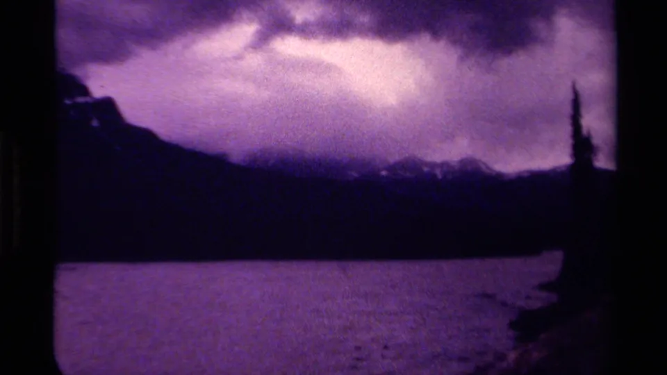 Purple-cast 8mm view of a Canadian lake, shadowed mountain ridge, clouded sky, and a dark tree shape at frame edge.