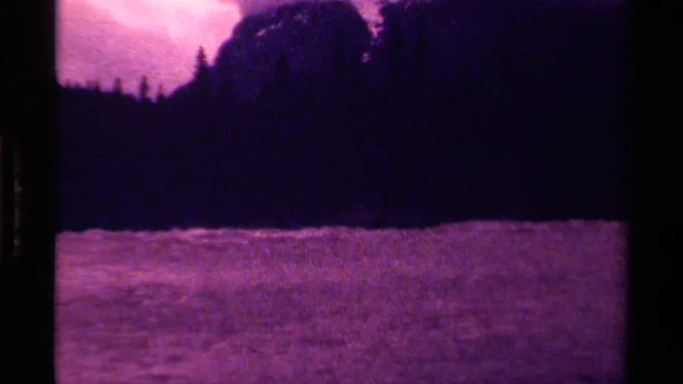 Late-frame 8mm view of a Canadian lake with dark forested shore, mountain silhouette, and magenta-purple film cast.
