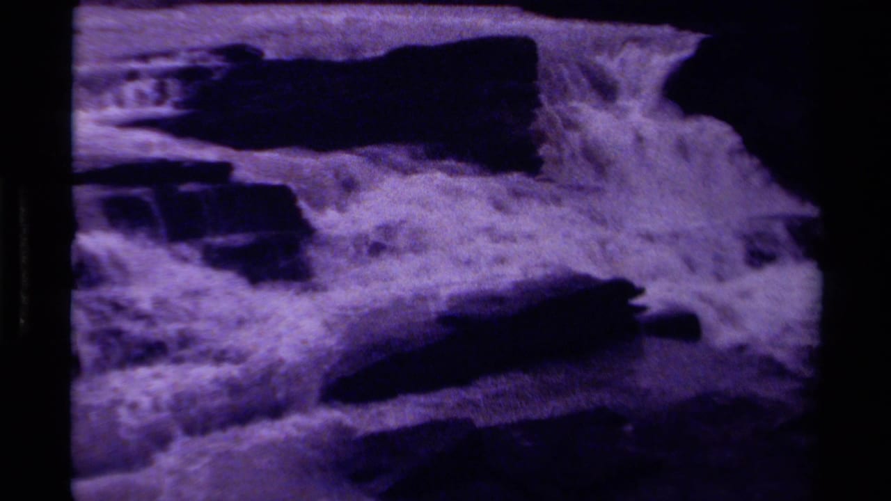 Foaming waterfall rushing over dark rock ledges with purple-tinted film grain.