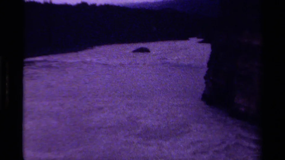 1979 Canadian 8mm frame showing fast, rippled water sweeping along a dark rocky shoreline, softened by grain and low exposure.