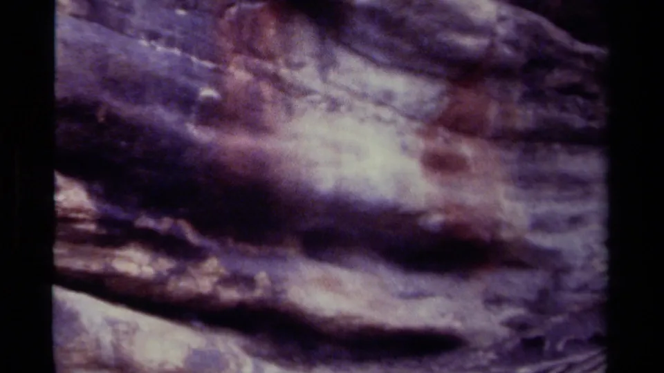 Close 8mm view of an indistinct rock face, showing muted tan highlights, purple shadows, grain, and handheld blur.