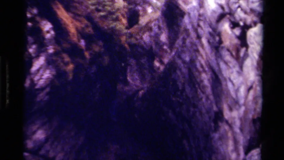 1979 Canadian home-movie frame showing a shadowed cave passage, rough rock surfaces, and a small reddish figure partly obscured.