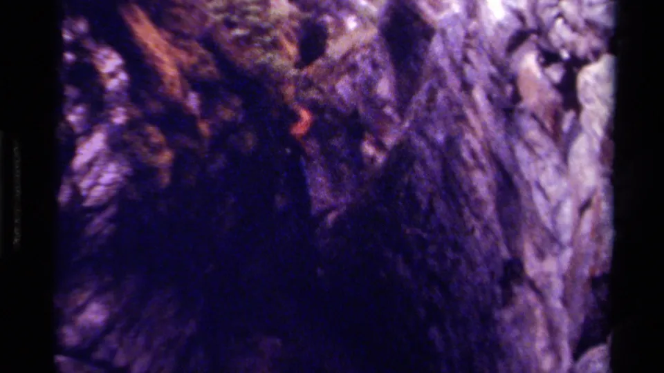 Grainy 1979 Canada 8mm frame of a person in reddish clothing moving through a narrow cave passage beside textured rock.
