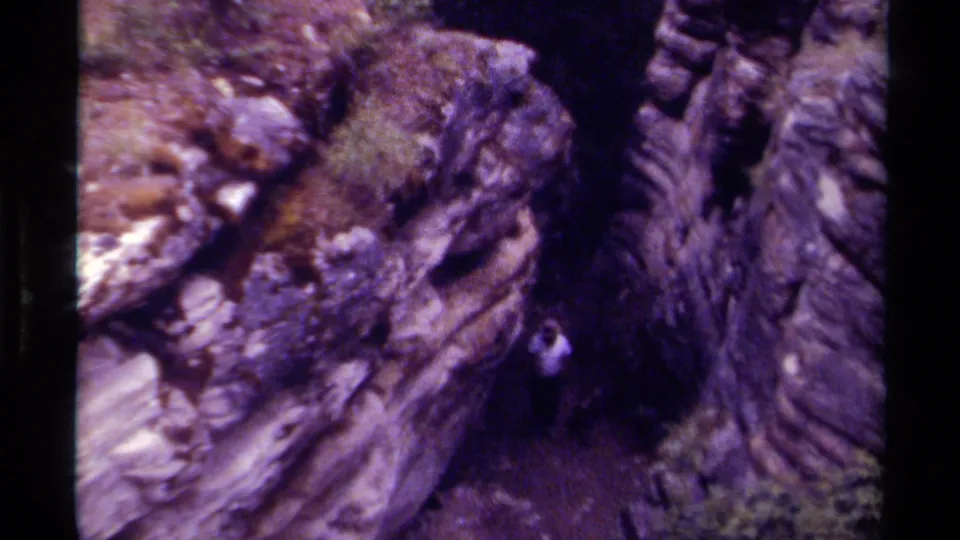 1979 Canadian 8mm frame of a hiker partly visible near dark, moss-covered rock formations in a wooded gorge.
