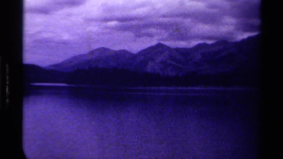 1979 Canadian 8mm frame of a calm lake with distant mountain peaks, a dark tree line, and heavy cloud cover.