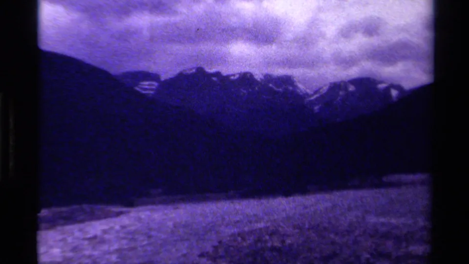 Purple-tinted 1979 Canadian home-movie view of mountains under a heavy sky, with a pale river or shoreline band below.