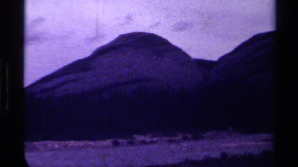 1979 Canadian 8mm frame with rounded hills, heavy shadow, bright foreground ground or water, and a cloudy sky.