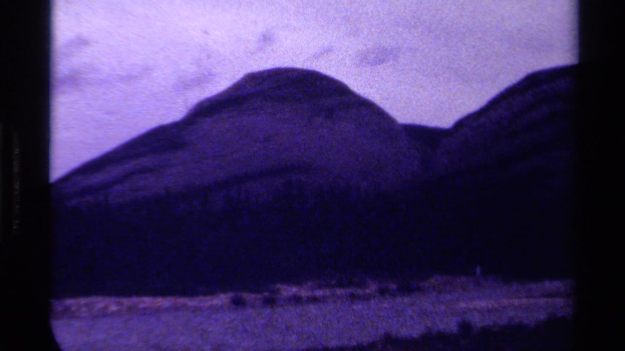 1979 Canada 8mm home-movie frame showing distant rounded mountains, dark forest, pale valley, grain, and purple color fading.