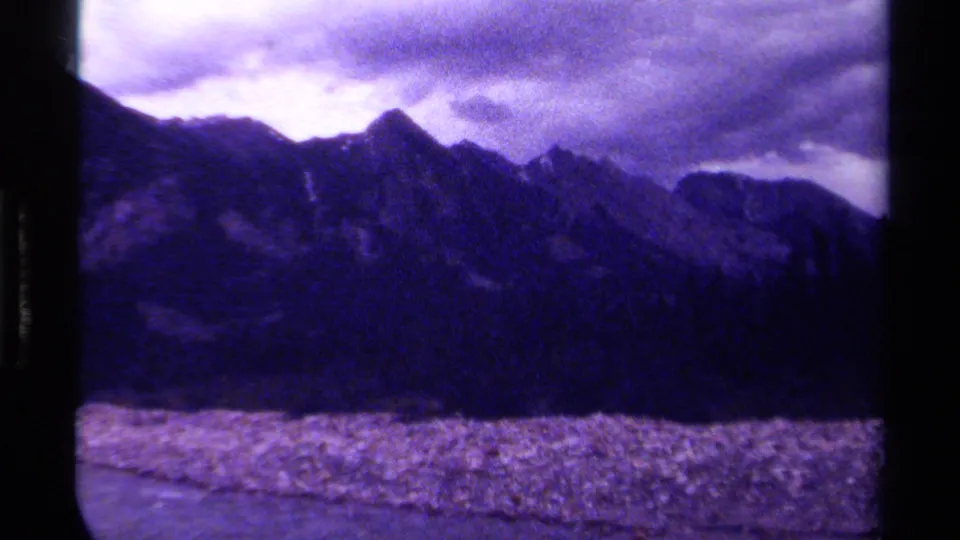 1979 Canada home-movie frame showing jagged mountain peaks, cloudy sky, dark forest, pale foreground, soft focus, and purple film tint.