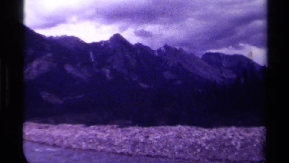 1979 Canada 8mm travel frame with mountain peaks under cloud cover, dark tree silhouettes, pale valley foreground, and visible film grain.