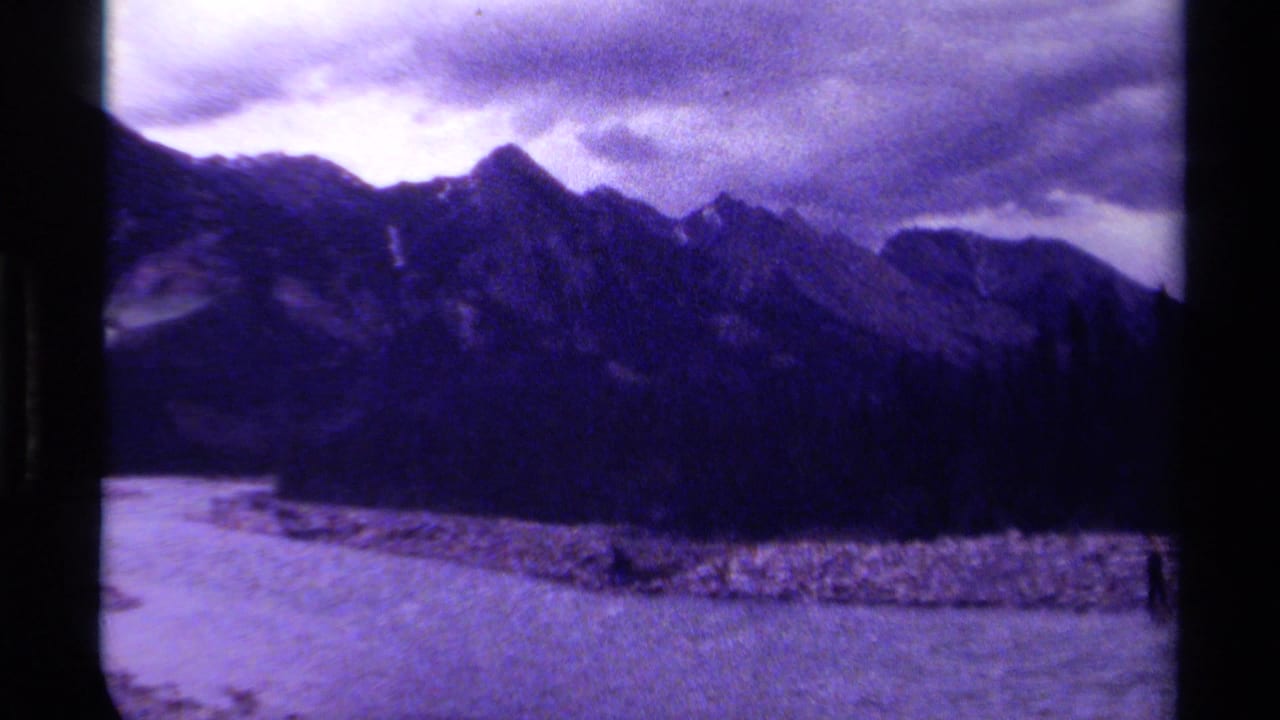 1979 Canadian 8mm frame showing dark mountain peaks, water, rocky shoreline, and overcast sky with a strong purple cast.