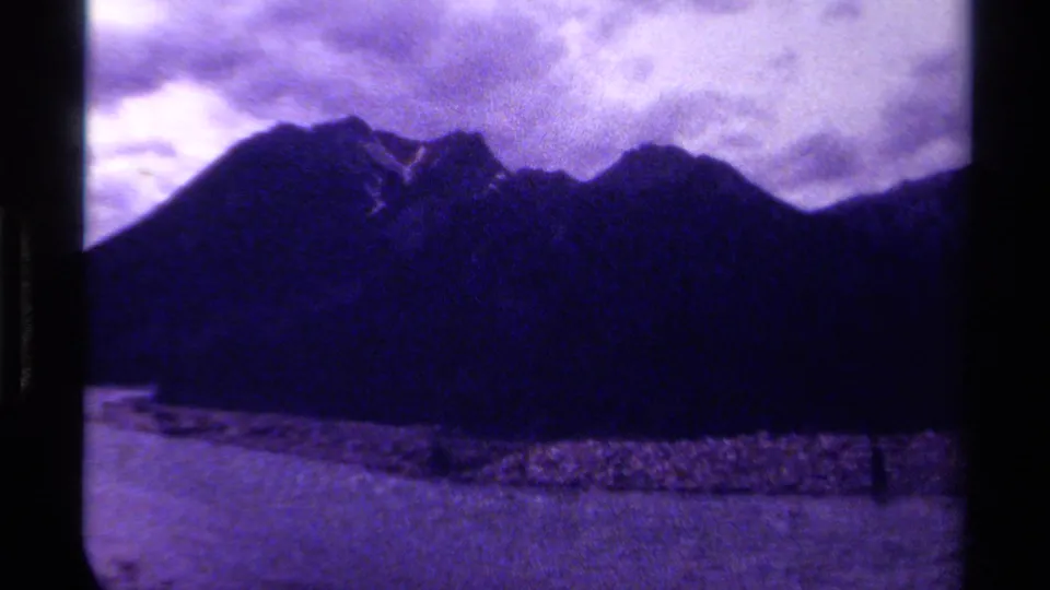1979 Canadian 8mm view of a shadowed mountain range across water, with rocky shoreline and cloud cover tinted purple-blue.