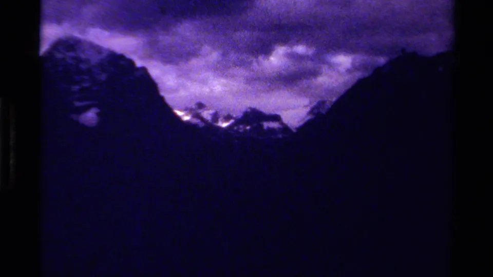 1979 Canadian 8mm frame with dark ridgeline silhouettes, small snow-bright peaks, and turbulent purple-blue cloud cover.