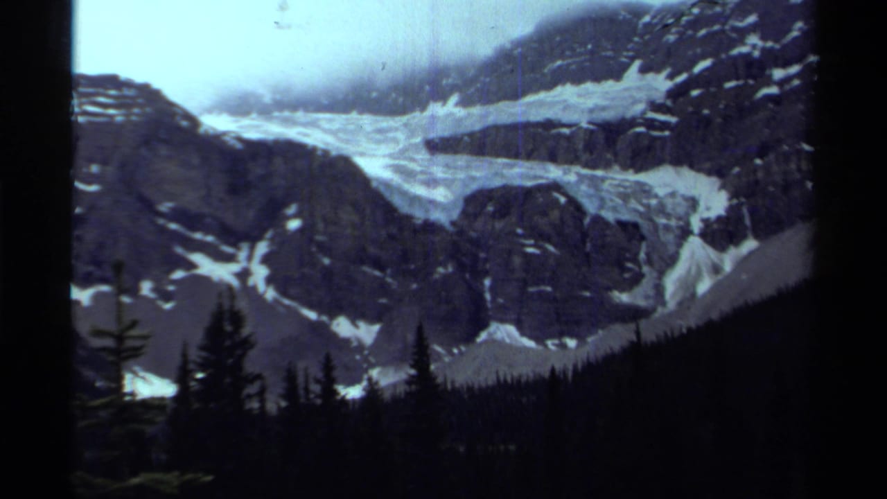 1979 Canadian 8mm frame showing a glacier winding through rocky peaks above silhouetted evergreens.