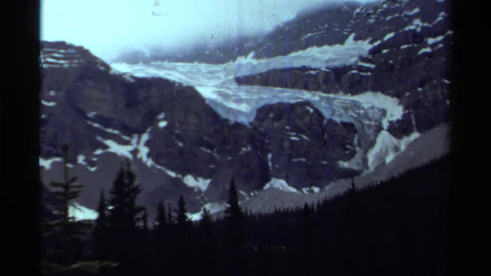 1979 Canadian 8mm frame showing a glacier winding through rocky peaks above silhouetted evergreens.
